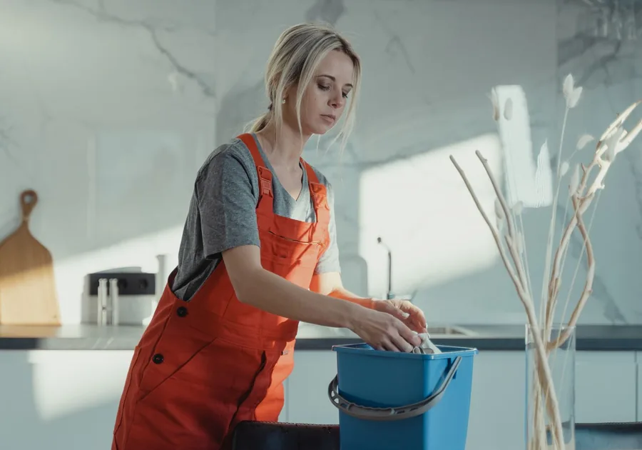 Intervenante en tenue orange effectuant le ménage dans une cuisine lumineuse — AXEO Services à Manosque